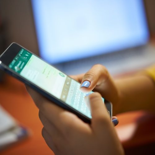 Young woman using cell phone to send text message on social network at night. Closeup of hands with computer laptop in background